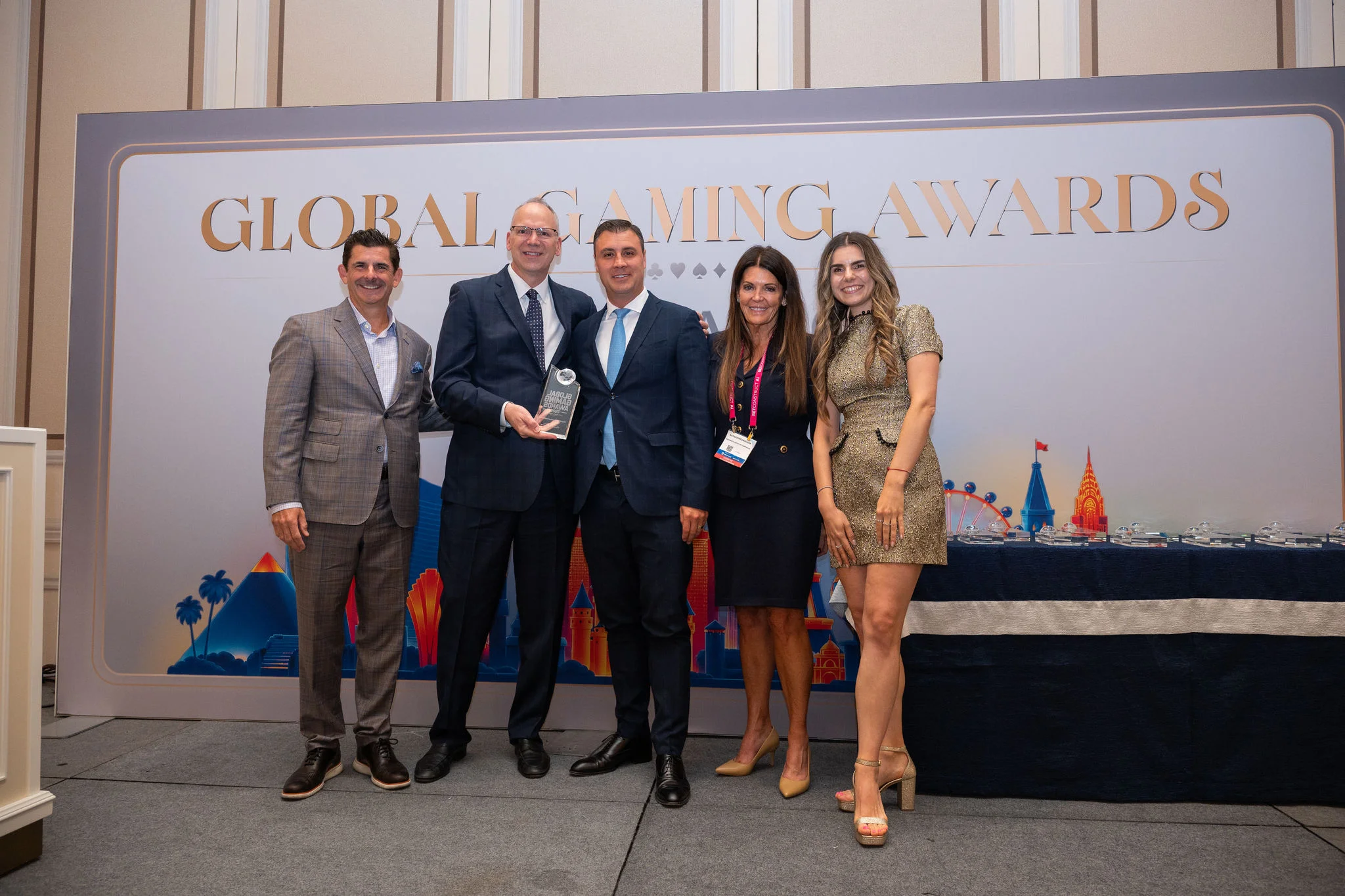Five adults stand together on a stage at the Global Gaming Awards event. One man in the center holds a trophy while the others smile beside him. The backdrop features the text "GLOBAL GAMING AWARDS" and colorful cityscape illustrations. The setting appear