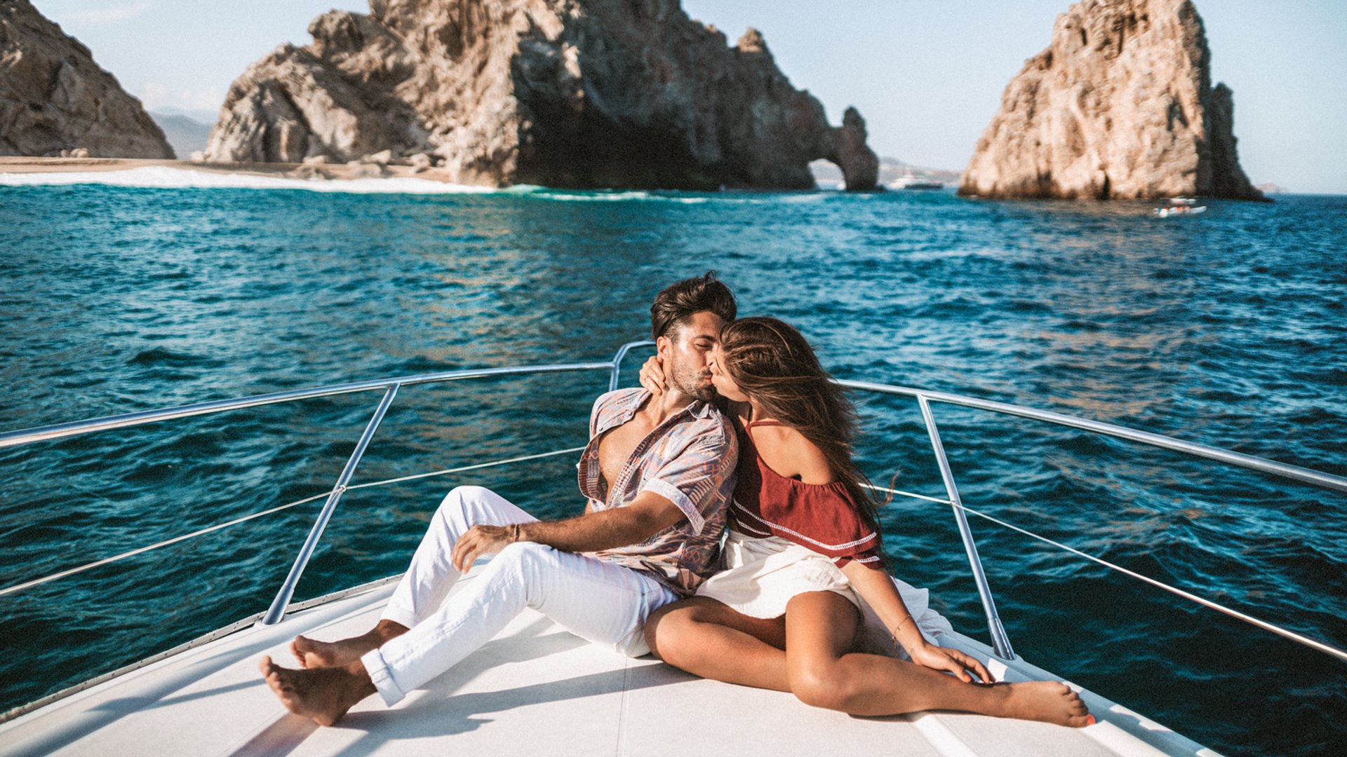 Couple kissing on a boat in front of El Arco rock formations, Los Cabos, with clear blue water and sky.