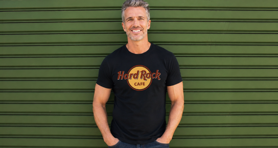 Smiling male model wearing a black Hard Rock Cafe logo T-shirt and jeans, with his hands in his pockets, against a green paneled wall.